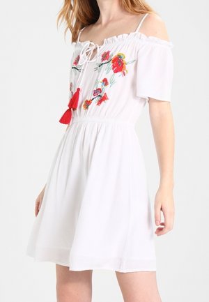 Biała sukienka z odkrytymi ramionami, czerwoną kwiatową haftowaną aplikacją i wiązaniami z frędzlami, krótkie rękawy i marszczona talia, noszona przez kobietę.