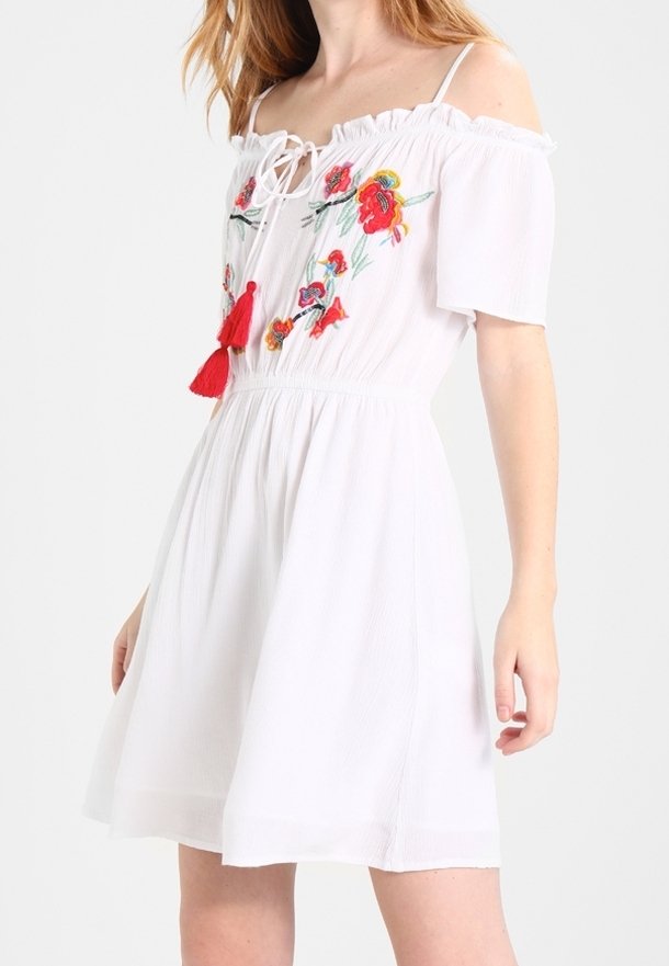 Robe blanche à épaules dénudées avec broderie florale rouge et liens à pompons, manches courtes et taille froncée portée par une femme.