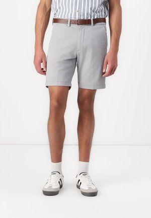 Mand iført lysegrå shorts, brun bælte, stribet skjorte, hvide sokker og hvide sneakers med sorte striber.