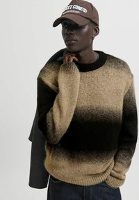 Person in einem braun-schwarz verlaufenden Strickpullover, dunklen Jeans und einer braunen Kappe mit weißem Text, die eine Jacke über der Schulter hält.