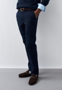 Hombre vistiendo pantalones azul marino, camisa azul claro con las mangas remangadas, cinturón marrón tejido y mocasines marrón oscuro, de pie con las manos en los bolsillos.