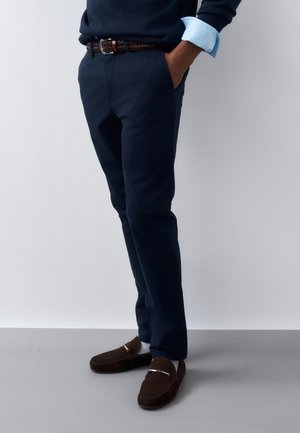 Άνδρας που φοράει ναυτικό παντελόνι, γαλάζιο πουκάμισο με μανίκια γυρισμένα, πλεκτή καφέ ζώνη και σκούρα καφέ μοκασίνια, στέκεται με τα χέρια στις τσέπες.