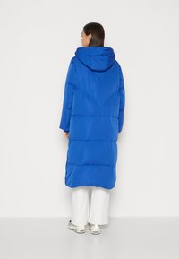 Blå polstret lang jakke med hette, med et quiltet design, en rett silhuett og trykknapper langs sidesømmen.