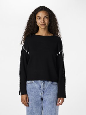 Woman with long curly hair wearing black sweater with white stitch details along sleeves and light blue jeans, standing against white background.