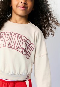 Hellbeige Cropped-Sweatshirt mit "HAPPINESS" in pinker Schrift, mit gerippten Bündchen und Saum. Kombiniert mit roten Hosen.