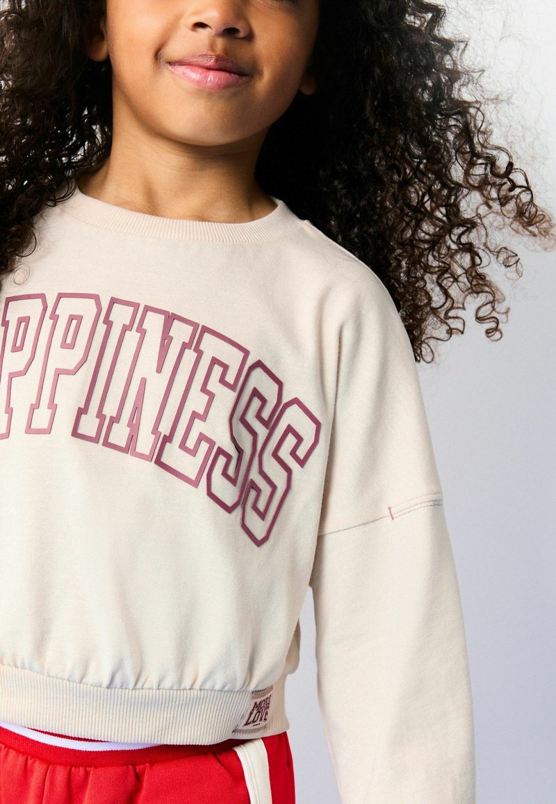 Hellbeige Cropped-Sweatshirt mit "HAPPINESS" in pinker Schrift, mit gerippten Bündchen und Saum. Kombiniert mit roten Hosen.