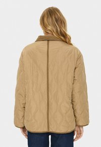 Tan quilted jacka med vågmönster, krage och kontrasterande kant. Har en avslappnad passform, långa ärmar och rundad fåll vid midjan.