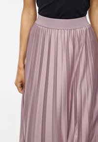 Záhybová sukňa v mäkkej ružovej látke, s širokým elastickým pásom a vertikálnymi záhybmi, ktoré vytvárajú textúru a prúd.