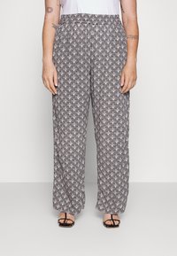 Vrouw in losse zwart-witte geometrische patroonbroeken en zwarte sandalen met straps, staande tegen een effen lichte achtergrond.