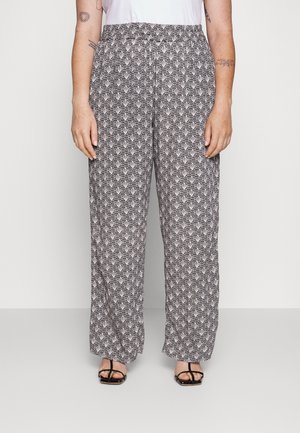 Vrouw in losse zwart-witte geometrische patroonbroeken en zwarte sandalen met straps, staande tegen een effen lichte achtergrond.