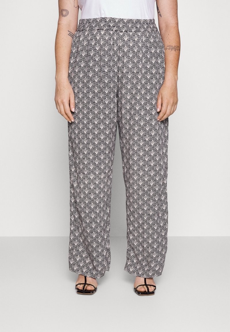 Vrouw in losse zwart-witte geometrische patroonbroeken en zwarte sandalen met straps, staande tegen een effen lichte achtergrond.