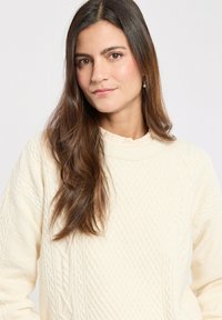 Maglione cream a trama cable con colletto alto e motivi testurizzati. Maniche lunghe e vestibilità rilassata, che mette in mostra un design a maglia grossa.