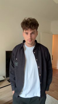 Cămașă neagră, cu nasturi, având un model texturat peste un tricou alb. Purtați cu pantaloni negri, afișând un look casual, în straturi.