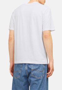 White cotton t-shirt with short sleeves and a crew neck, displayed from the back, paired with light blue denim jeans featuring subtle stitching.