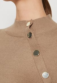 Gros plan d'un pull en maille côtelée beige avec des boutons-pression dorés sur le côté d'un col haut, porté par une personne aux cheveux bruns.