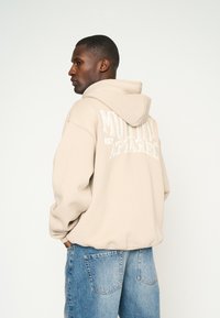 Sudadera beige con un gran gráfico en blanco de "MUTINY APPAREL" en la parte posterior. Cuenta con capucha con cordón y corte relajado, combinada con jeans de color azul claro.