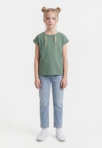 Niña joven con dos moños trenzados, vestida con camiseta verde de manga corta, vaqueros azules claros, calcetines blancos y zapatillas blancas, de pie frente a un fondo blanco.