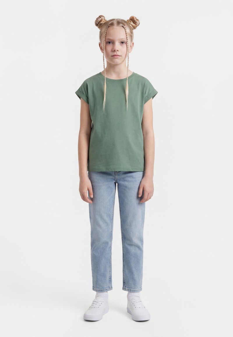 Niña joven con dos moños trenzados, vestida con camiseta verde de manga corta, vaqueros azules claros, calcetines blancos y zapatillas blancas, de pie frente a un fondo blanco.