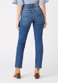 Jeans de denim azul con corte recto, que presentan una cintura alta y dos bolsillos traseros con costuras en zigzag. Combinados con zapatos de tacón color marrón claro.