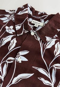 Braunes Kurzarmhemd mit weißen Blumenmustern, einem Kragen und zwei Knöpfen. Der Stoff wirkt strukturiert. Etikett sichtbar.