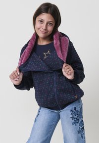 Ragazza che sorride, indossando una giacca blu navy con fodera in pile viola e fantasia floreale, e jeans azzurri chiari con ricami blu scuro.