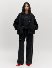 Zwarte oversized bomberjack met drukknoopsluitingen, gecombineerd met een zwarte top en wijde broek. Rode glanzende schoenen maken de outfit af.