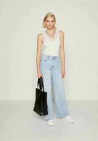 Jovem mulher a usar um top de alças branco com rendas, calças largas azul claro, sapatos brancos, segurando uma mala tote preta.