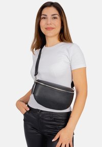 Schwarze Leder-Crossbody-Tasche mit strukturiertem Finish, Reißverschluss, und verstellbarem Tragegurt, um die Taille getragen, kombiniert mit einem weißen Hemd.