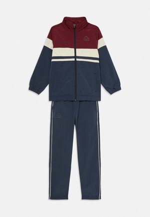 Survêtement avec un haut bordeaux, un bas marine, présentant des rayures blanches et un logo. Fabriqué en matériau léger avec des poignets et une taille élastiques.