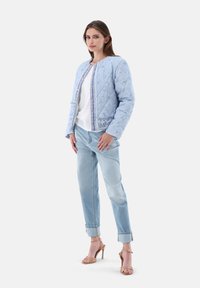 Helleblaue gesteppte Jacke mit Paisleymuster, kombiniert mit einem weißen Oberteil und hellblauen Jeans. Das Modell trägt beigefarbene High-Heels.