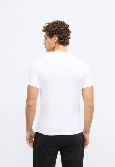 Balts īss piedurkņu T-krekls ar apaļu apkakli, gluda tekstūra, pieguļošs dizains. Modelis to valkā kopā ar melnām biksēm, skatoties prom no kameras.