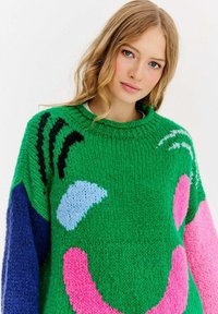 Grüner Strickpullover mit auffälligen Mustern: rosa und hellblaue Formen, schwarze Streifen und blaue Akzente an den Ärmeln. Weicher, strukturierter Stoff.