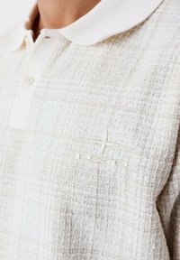 Beige poloshirt met een textuurpatroon, voorzien van een witte kraag en een subtiele geborduurde logo op de borst.