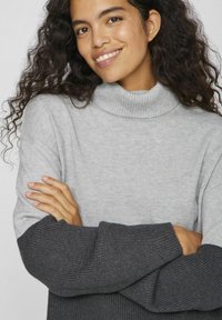 Jersey de cuello alto gris claro con mangas acanaladas gris oscuro. Textura suave y ajuste relajado. La modelo está con los brazos cruzados y sonríe.