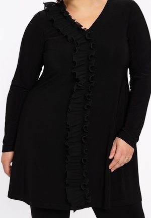 Robe noire à manches longues avec un galon froncé vertical sur le devant, portée par une personne portant une bague à la main droite.