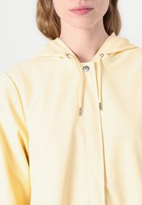 Chubasquero amarillo con capucha, cierre frontal con botón a presión y cordones ajustables. Textura suave, material ligero y diseño casual.