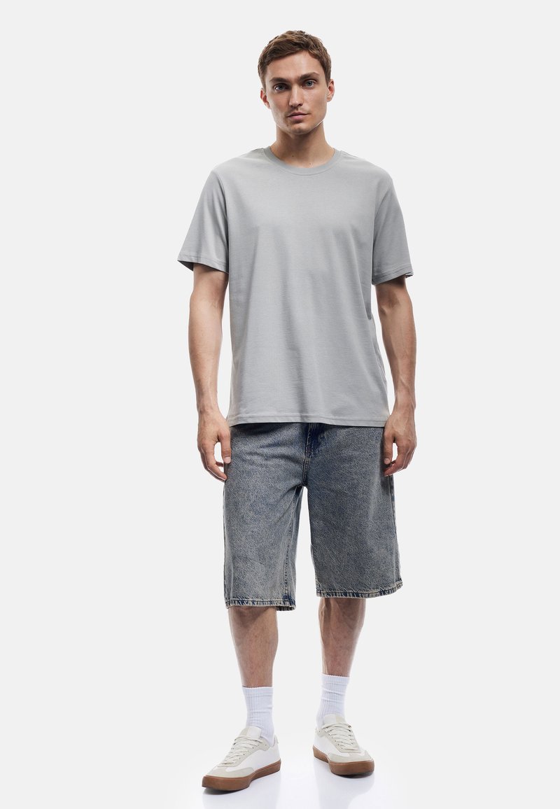 Camiseta de manga corta gris claro combinada con pantalones cortos de mezclilla, calcetines blancos y zapatillas claras. Diseño sencillo, corte relajado, detalles mínimos.