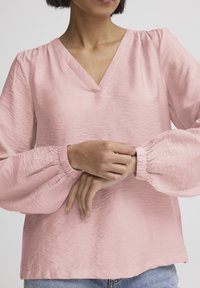 Světle růžová halenka s výstřihem do V, nadýchanými dlouhými rukávy, texturovanou látkou a nařasenými manžetami, doplněná modrými džínami z denimu.