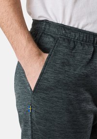 Dark grey athletic pants with a textured finish, featuring an elastic waistband and side pockets. Includes a small, blue and yellow flag emblem.