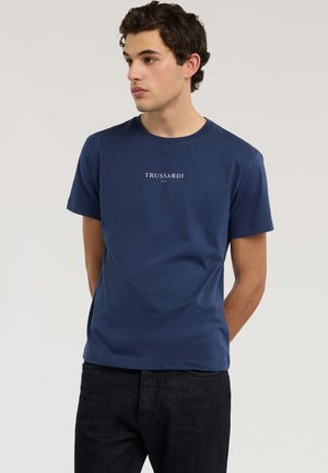 Młody mężczyzna o kręconych ciemnych włosach, ubrany w granatową koszulkę z logo Trussardi i czarne spodnie, patrzący w lewo z rękami za plecami.
