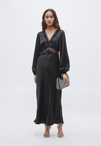 Čierne saténové šaty s čipkovým lemom, s dlhými rukávmi a voľnou siluetou. Dopĺňajú ich trblietavé psaníčko a sandále s remienkami.