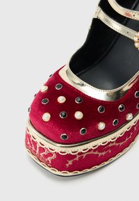 Παπούτσια πλατφόρμας από κόκκινο βελούδο με χρυσές λεπτομέρειες, διακοσμημένα με μαύρες και μαργαριταρένιες τρούκ, και μια διακοσμητική, κυματιστή τρέσα στη βάση.