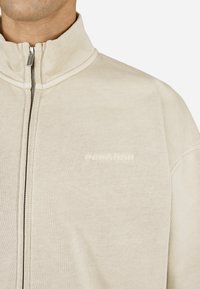 Bežs zip-up jaka, izgatavota no mīksta auduma ar augstu apkakli. Iespējamās tonālās izšūtās emblēmas uz krūtīm. Vienkāršs dizains, brīvs piegriezums.