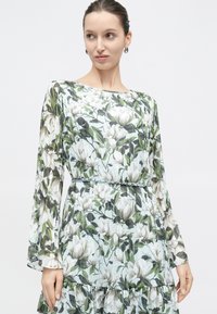 Vrouw draagt een lange mouw bloemenjurk met groene bladeren en witte bloemen, getailleerd met een zilveren kettingriem.