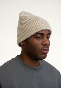 Pletená béžová čepice s špičatým vrchem a jemnou žebrovanou texturou, nošená přes šedou mikinu s kulatým výstřihem. Obsahuje vyšívané logo.