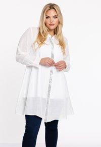 Witte linnen tuniek met lange mouwen en een knoopsluiting aan de voorkant. Heeft een zilveren metallic strook in het midden. Gepaarde het met donkere denim broeken.
