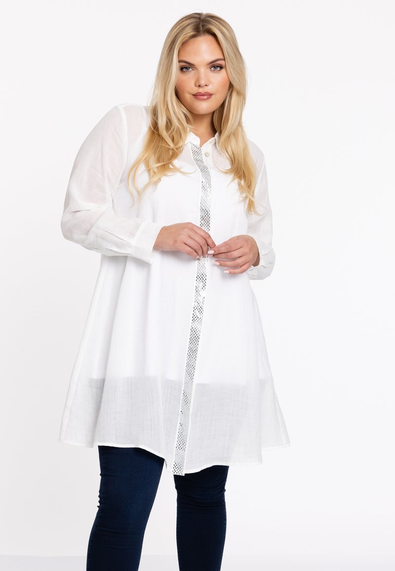 Witte linnen tuniek met lange mouwen en een knoopsluiting aan de voorkant. Heeft een zilveren metallic strook in het midden. Gepaarde het met donkere denim broeken.