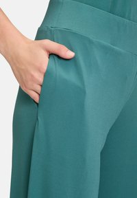 Teal trousers with a smooth texture, featuring a side pocket and a soft waistband, highlighting a tailored, wide-leg design.