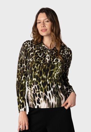 Kvinde med langt hår iført en langærmet, grøn og sort leopardprint skjorte og sorte bukser, poserer foran en ensfarvet baggrund.