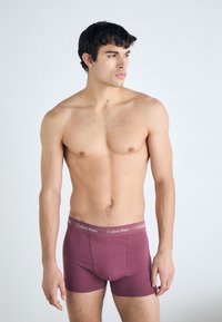 Calvin Klein maroon boxer briefs feature a logo waistband, fitted design, and soft cotton material, displayed on a model with no shirt.